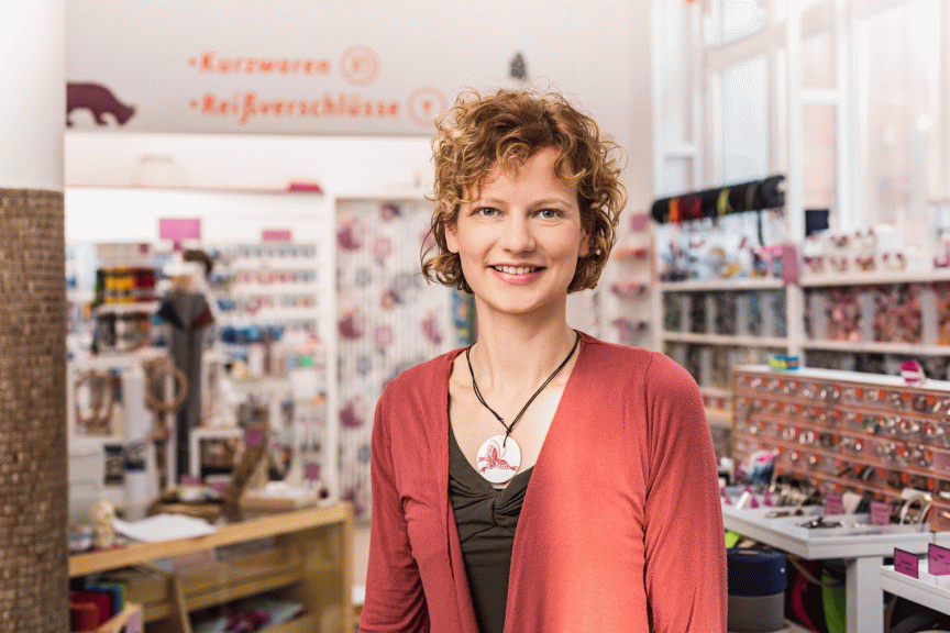 Frau mit lockigem Haar und roter Strickjacke steht lächelnd in einem Kurzwaren- und Reißverschluss-Geschäft.