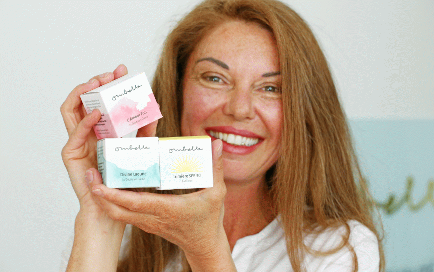 Lächelnde Frau hält drei kleine Kosmetikprodukte der Marke "Ombelle" in die Kamera, darunter eine SPF 30 Creme.