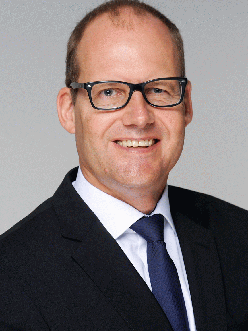 Mann mit Brille, kurzem Haar, Anzug und Krawatte lächelt vor grauem Hintergrund. Professionelles Porträtfoto.