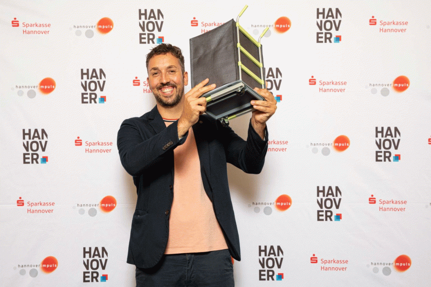 Mann hält lächelnd einen schwarzen Preis/Award in die Höhe vor einer Hannover-Sparkasse-Sponsorenwand.