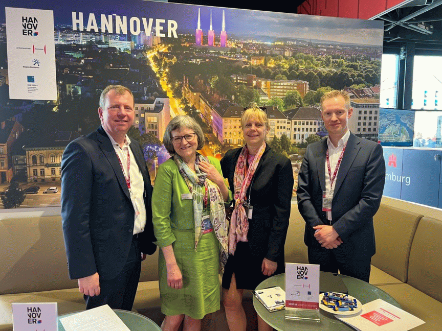Vier Personen stehen vor einem Hannover-Stadtpanorama-Banner auf einer Messe. Alle tragen Namensschilder.