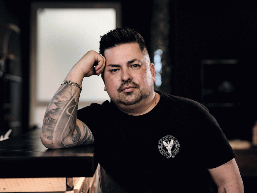Mann mit Tattoos am Arm, schwarzem T-Shirt und kurzem Haar, lehnt mit Hand am Kopf an einem Tisch.