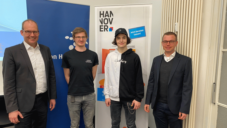 Vier Männer stehen vor einem Hannover-Banner. Zwei tragen Anzüge, zwei Jüngere sind casual gekleidet.