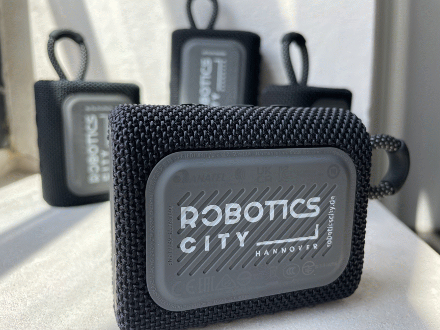 Drei schwarze Bluetooth-Lautsprecher mit Schlaufe und grauem Logo "Robotics City Hannover" auf rauer Textilhülle.