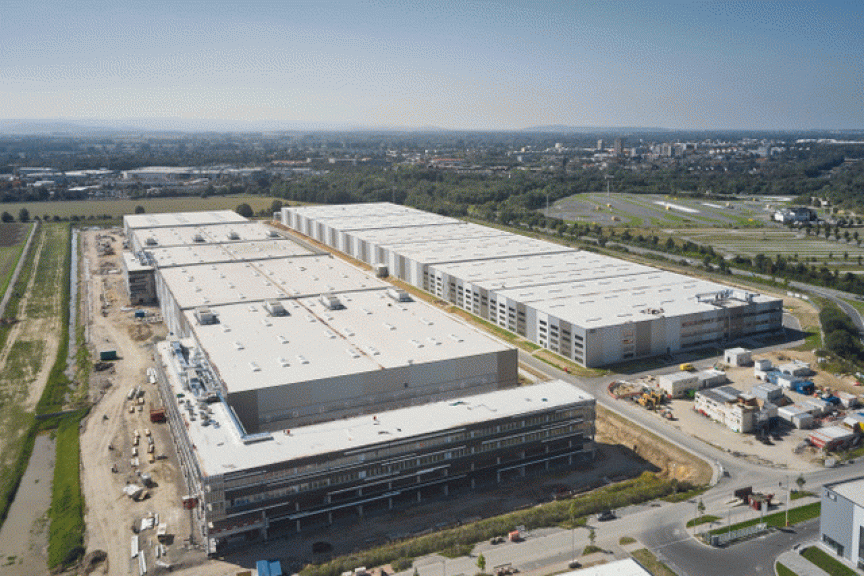 Luftaufnahme eines großen Industrie- oder Logistikkomplexes mit mehreren Flachdachgebäuden, umgeben von Baustellen und Grünflächen.