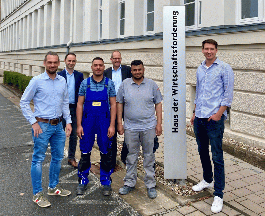 Sechs Männer stehen vor dem Schild „Haus der Wirtschaftsförderung". Zwei tragen Arbeitskleidung, die anderen Freizeitkleidung.