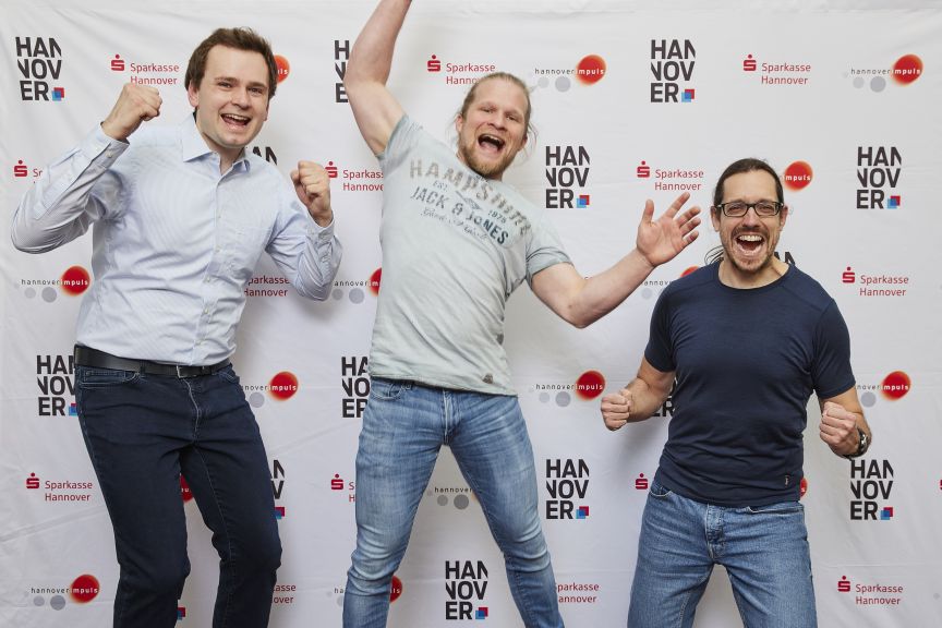 Drei Männer posieren jubelnd vor einem Hannover-Impulse- und Sparkasse-Hannover-Rollup-Banner.