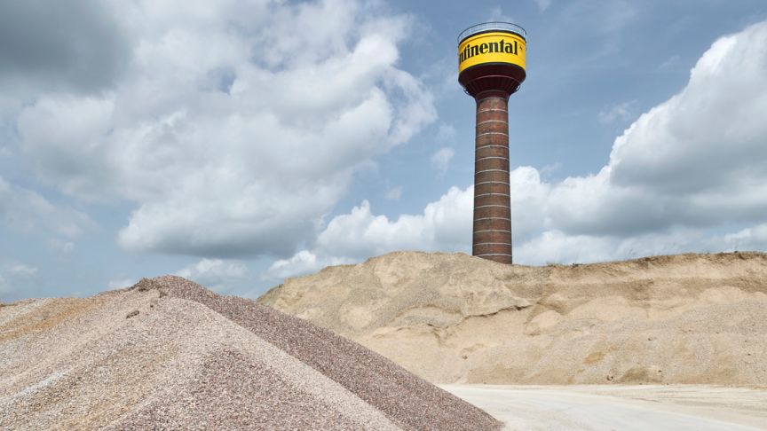 Wasserturm mit gelbem Continental-Logo hinter Schotterhaufen und Sandhügeln unter bewölktem Himmel.