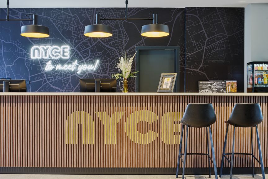 Hotelrezeption mit Holzverkleidung, „NYCE"-Logo, Neonschrift, zwei schwarzen Barhockern und Stadtkartenillustration.