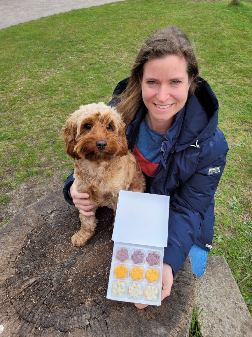 Frau mit blauer Jacke hält Schachtel mit pfotenfömigen Hundekeksen, neben ihr sitzt ein hellbrauner Cockerspaniel auf einem Baumstumpf.