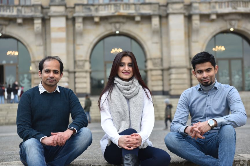 Drei junge Personen sitzen nebeneinander vor einem historischen Gebäude mit Bögen. Sie lächeln in die Kamera.