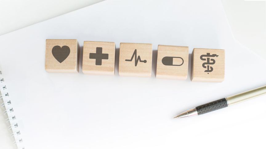 Fünf Holzwürfel mit medizinischen Symbolen (Herz, Kreuz, EKG, Kapsel, Äskulapstab) auf einem Notizbuch neben einem Stift.