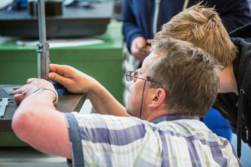 Ein Mann mit Brille misst mit einem Messschieber an einem Werkstück. Ein jüngerer Mann beobachtet ihn dabei.