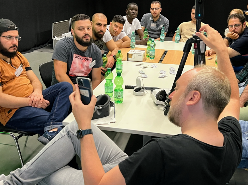 Gruppe junger Männer sitzt um einen Tisch mit Wasserflaschen. Ein Mann fotografiert mit Smartphone und Stativ.