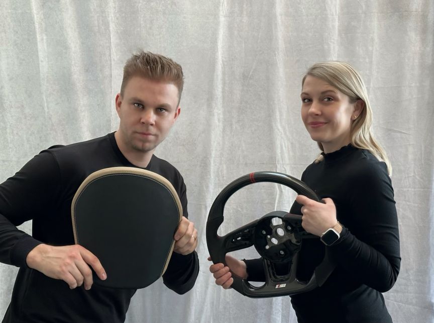 Zwei Personen in schwarzen Outfits halten je ein Lenkrad: links ein abgenutztes, rechts ein modernes Carbon-Lenkrad.