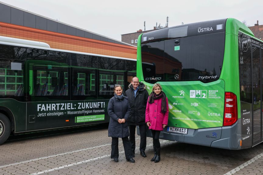 Drei Personen stehen vor zwei grünen ÜSTRA-Bussen. Aufschrift: „Fahrtziel: Zukunft! Wasserstoffregion Hannover."