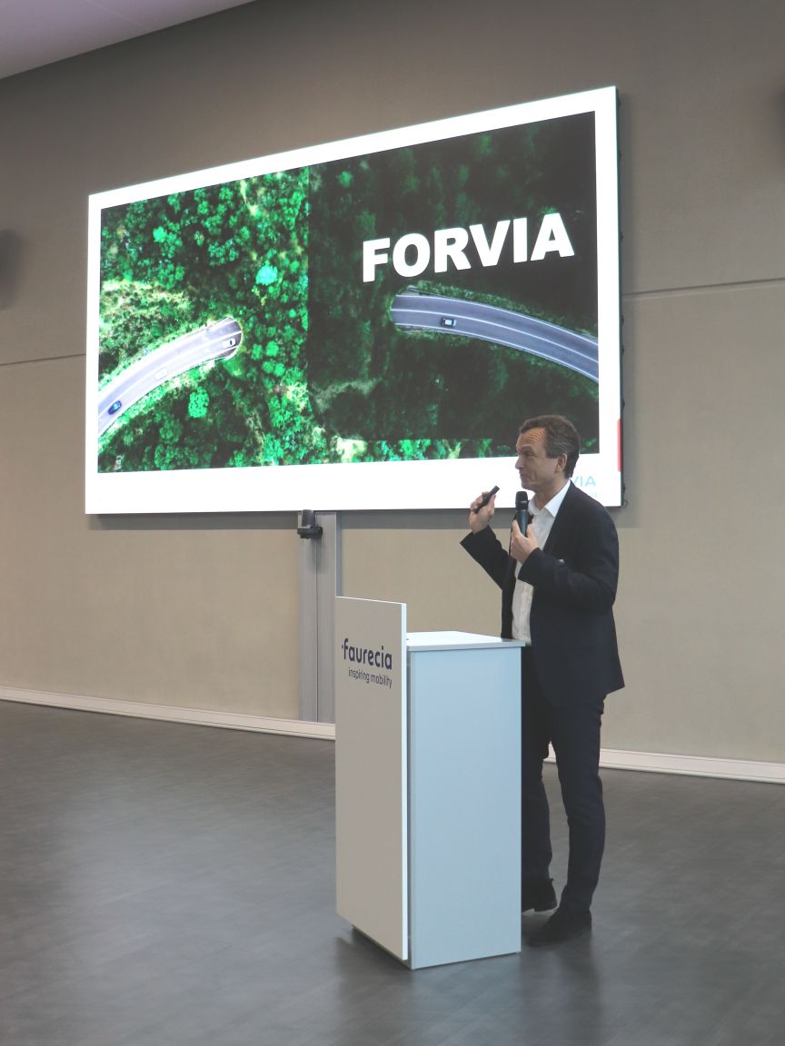 Mann im Anzug steht an Faurecia-Rednerpult mit Mikrofon, dahinter Leinwand mit "FORVIA"-Logo und Luftaufnahme einer Straße.
