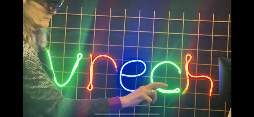 Person zeigt auf buntes Neonschild mit dem Wort „vrech" in Grün, Rot und Blau vor einem Gitterhintergrund.