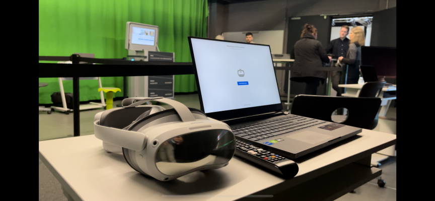 VR-Headset und Laptop auf einem Tisch, im Hintergrund Personen vor einem Greenscreen.