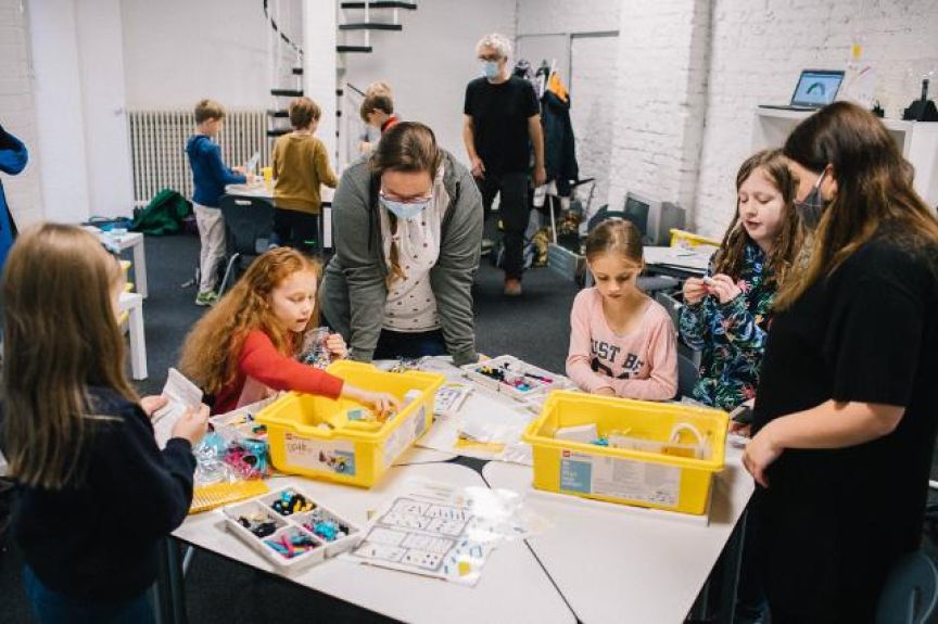 Kinder und Erwachsene mit Masken arbeiten an einem Tisch mit gelben Kisten voller Bausteine in einem hellen Raum.