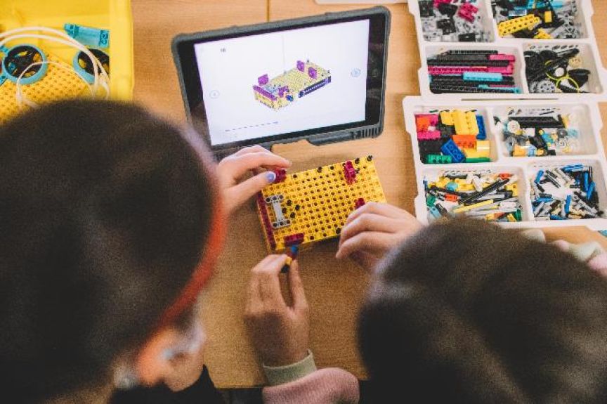 Zwei Kinder bauen mit gelben LEGO-Steinen, daneben ein Tablet mit 3D-Modell und Sortierbox mit bunten Bausteinen.