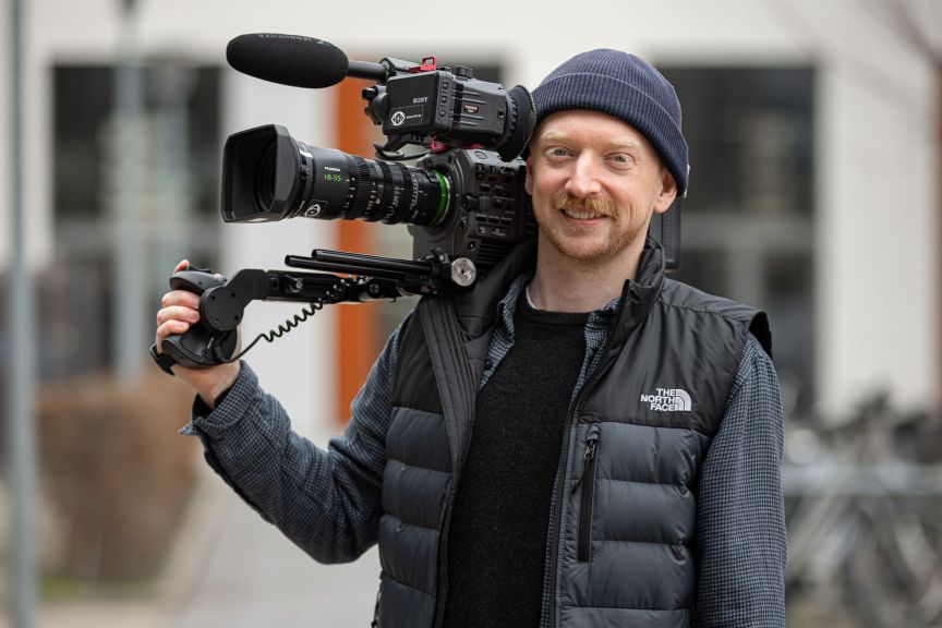 Mann mit blauer Mütze und North Face-Weste trägt eine Sony-Profikamera auf der Schulter.