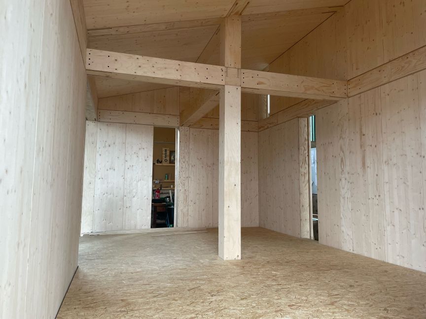 Leerer Innenraum mit Holzwänden, Holzdecke und OSB-Boden. Mittig ein Holzpfeiler, im Hintergrund Türöffnungen.