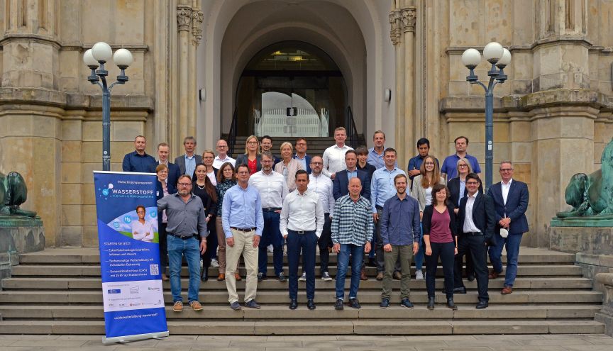 Gruppenfoto von ca. 30 Personen auf Treppe eines Gebäudes, daneben ein Banner mit Aufschrift „Wasserstoff".