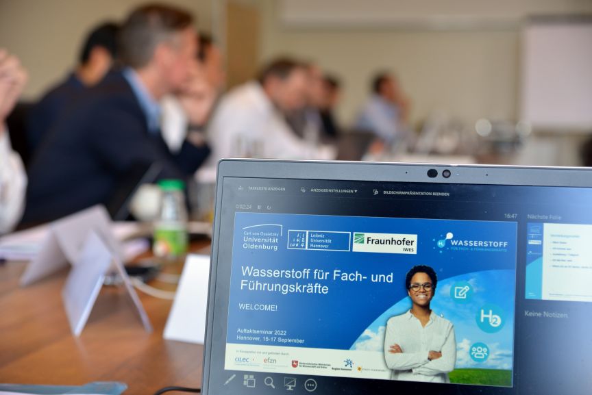 Laptop mit Präsentation „Wasserstoff für Fach- und Führungskräfte" im Vordergrund, Teilnehmer eines Seminars im Hintergrund.