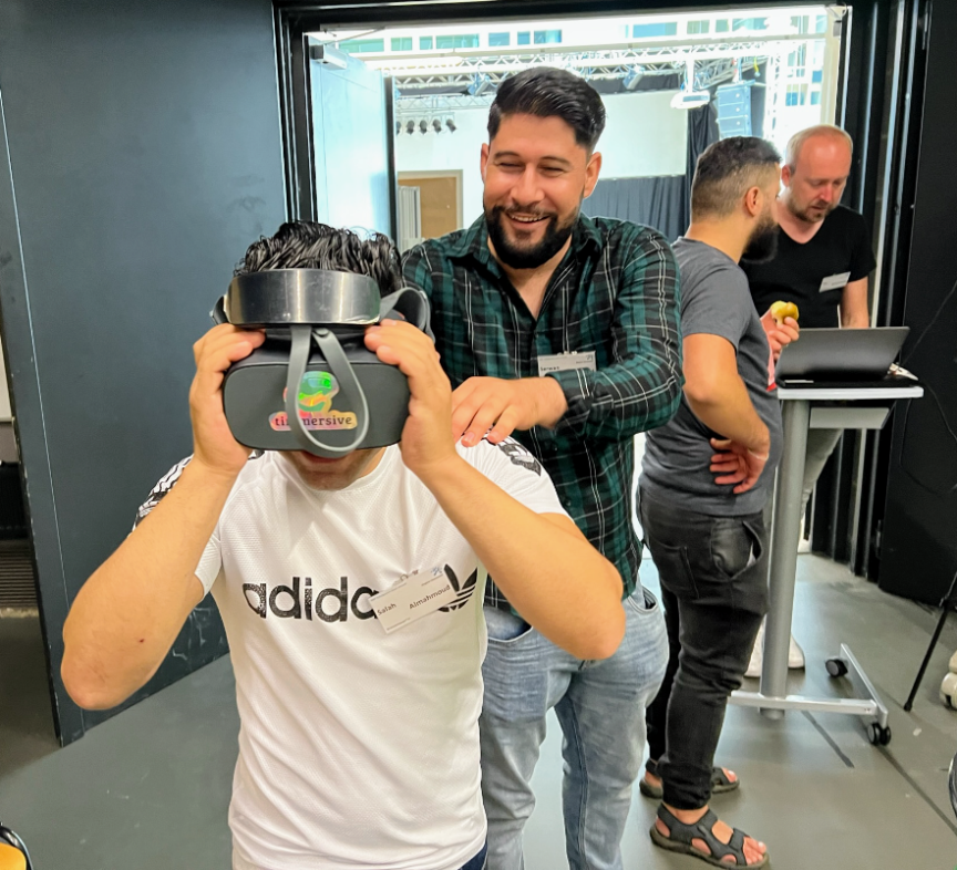 Mann mit Adidas-Shirt trägt VR-Brille, daneben stehender Mann in kariertem Hemd lächelt. Im Hintergrund zwei weitere Personen.