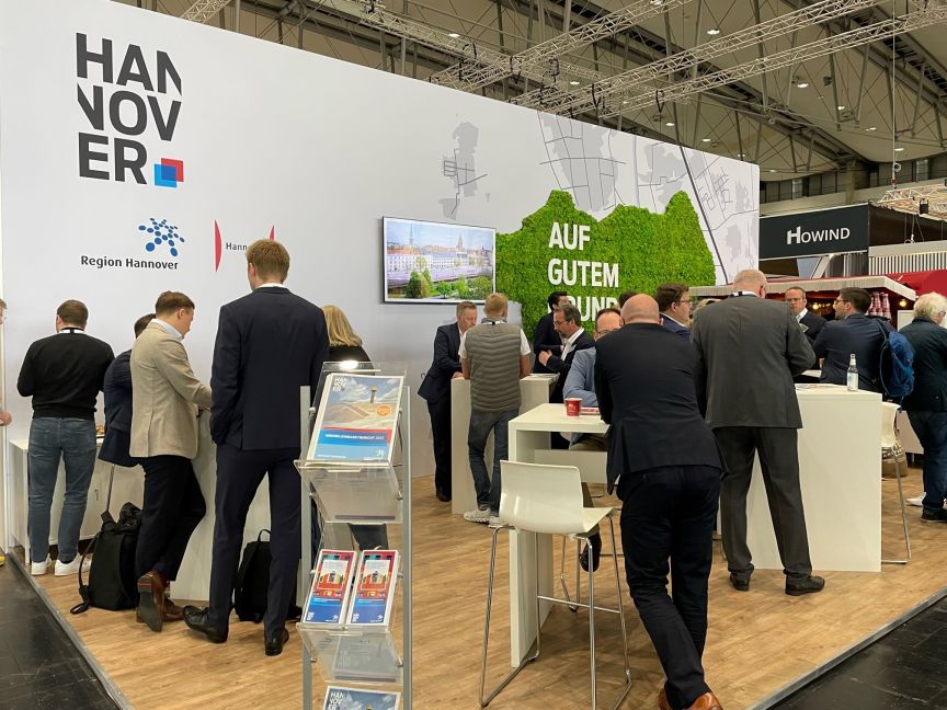 Messestand der Region Hannover mit weißen Stehtischen, Besuchern in Businesskleidung und grüner Pflanzenwand mit Slogan.