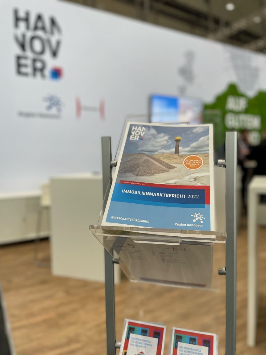 Prospektständer mit „Immobilienmarktbericht 2022" der Region Hannover vor einem Hannover-Messestand.