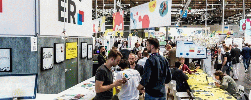 Messestand mit Besuchern in großer Halle, Schilder mit "Startup City" und bunten Logos, Personen stehen und sitzen an Tischen.