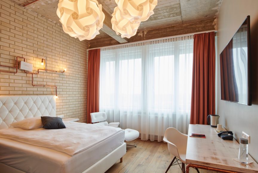 Hotelzimmer mit weißem Doppelbett, Ziegelwand, roten Vorhängen, Schreibtisch, TV und zwei Pendelleuchten.