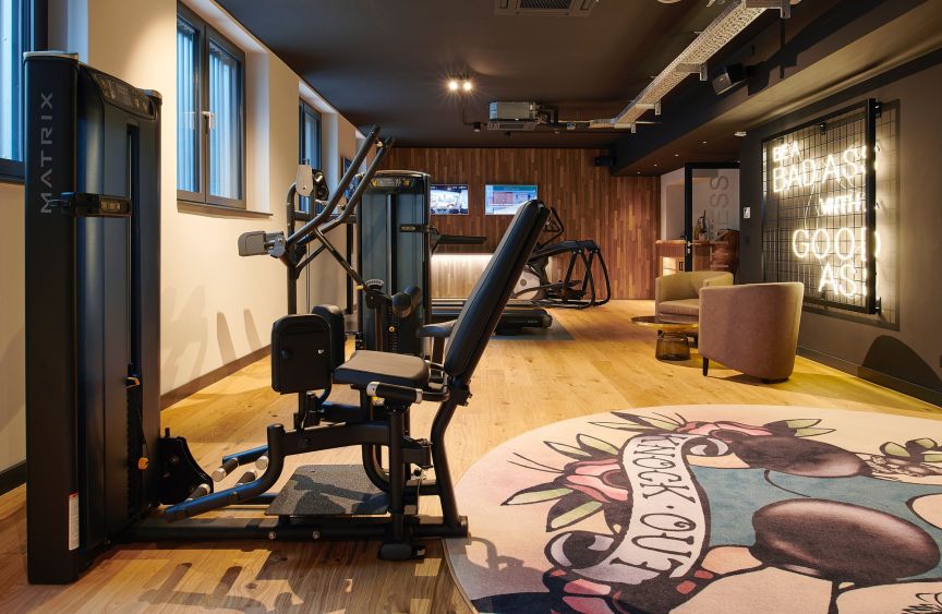 Fitnessraum mit schwarzen Kraftgeräten (Matrix), Holzboden, rundem Teppich, Neonschrift und Loungesessel.