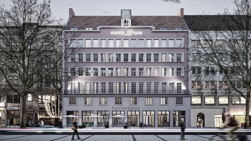 Mehrstöckiges Stadtgebäude „Motel One Kurierhaus" bei Dämmerung, beleuchtet, mit Passanten und kahlen Bäumen.