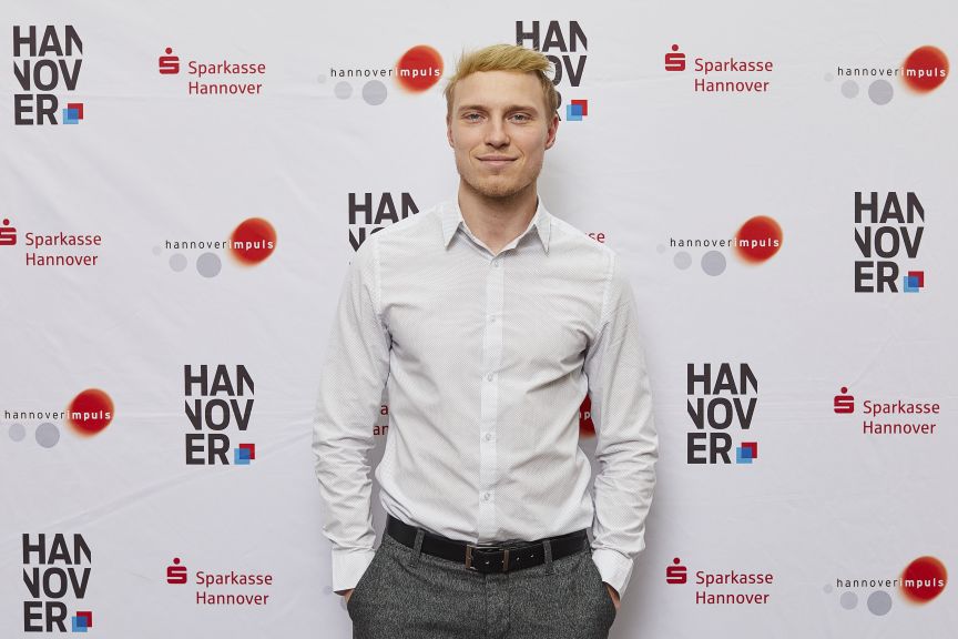 Junger Mann mit blonden Haaren in weißem Hemd und grauer Hose vor Hannover-Sponsorenwand.