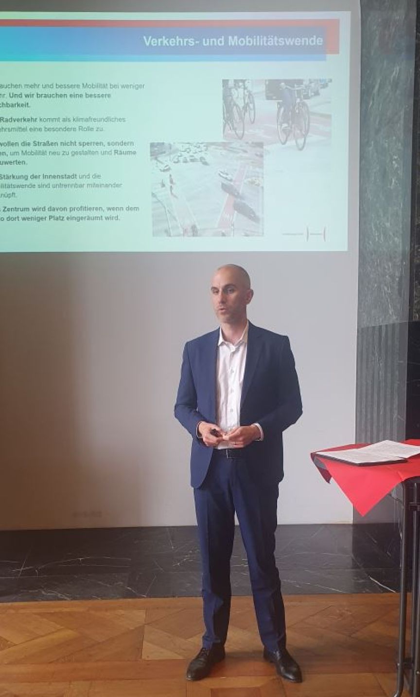Mann im blauen Anzug steht vor einer Präsentationsfolie mit der Aufschrift „Verkehrs- und Mobilitätswende".