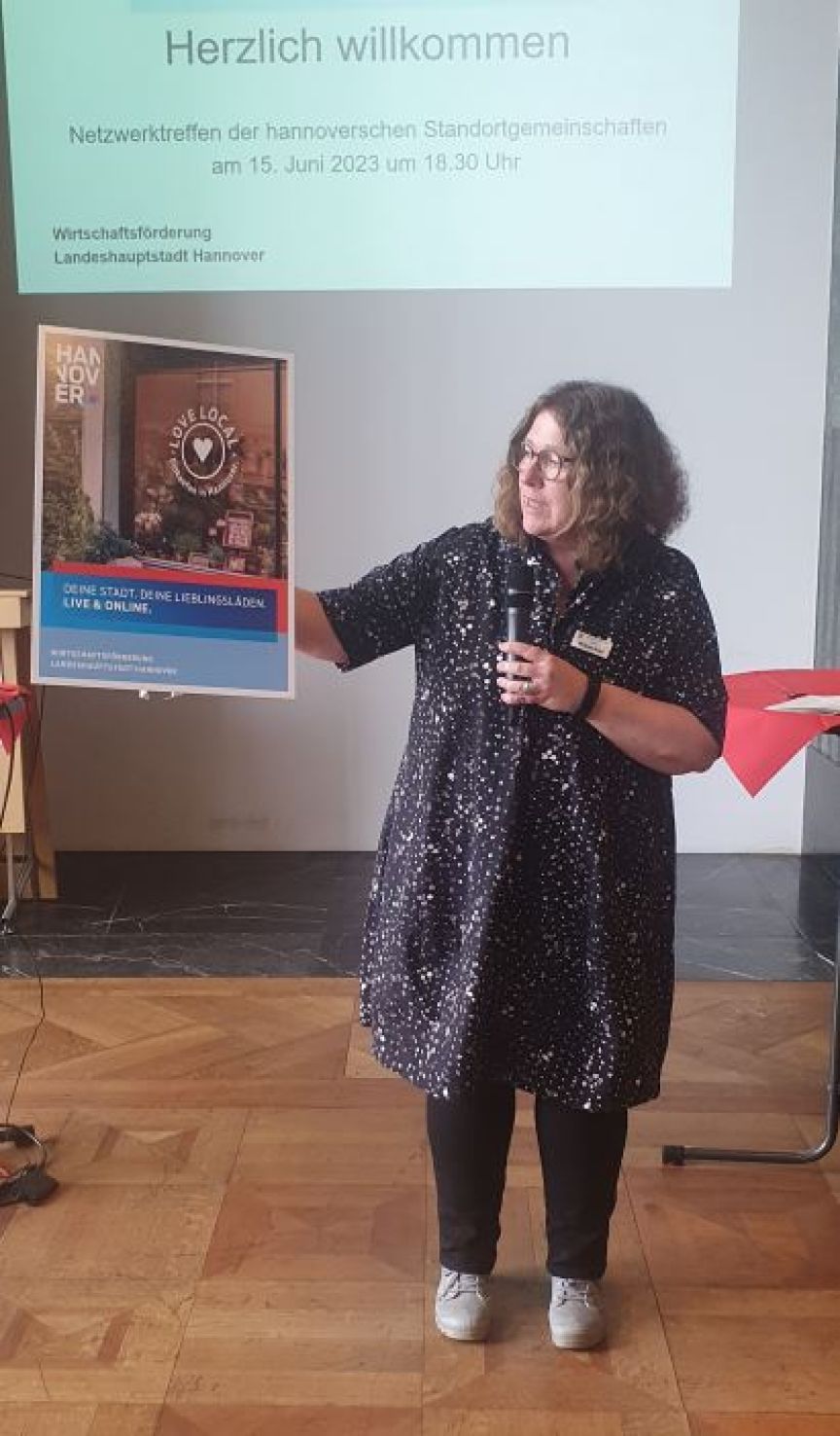 Frau hält Mikrofon und Schild auf Veranstaltung in Hannover, 15. Juni 2023, Projektionswand mit „Herzlich willkommen" im Hintergrund.