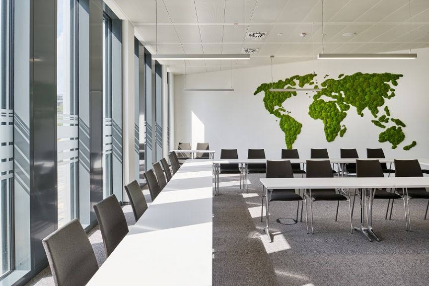 Konferenzraum mit weißen Tischen, grauen Stühlen, Glasfronten und einer Weltkarte aus Moos an der Wand.