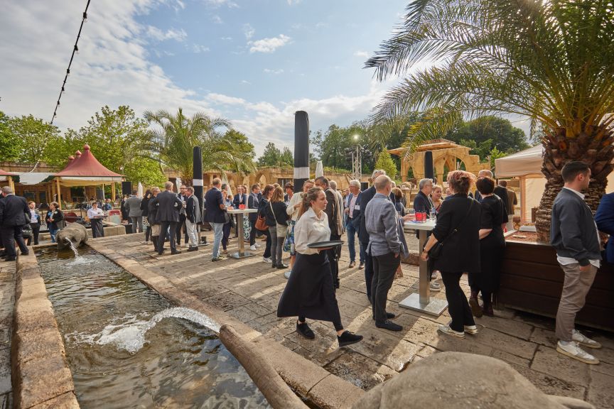 Outdoor-Veranstaltung mit vielen Menschen, Palmen, einem Wasserkanal, Stehtischen und einem Servicepersonal mit Tablett.