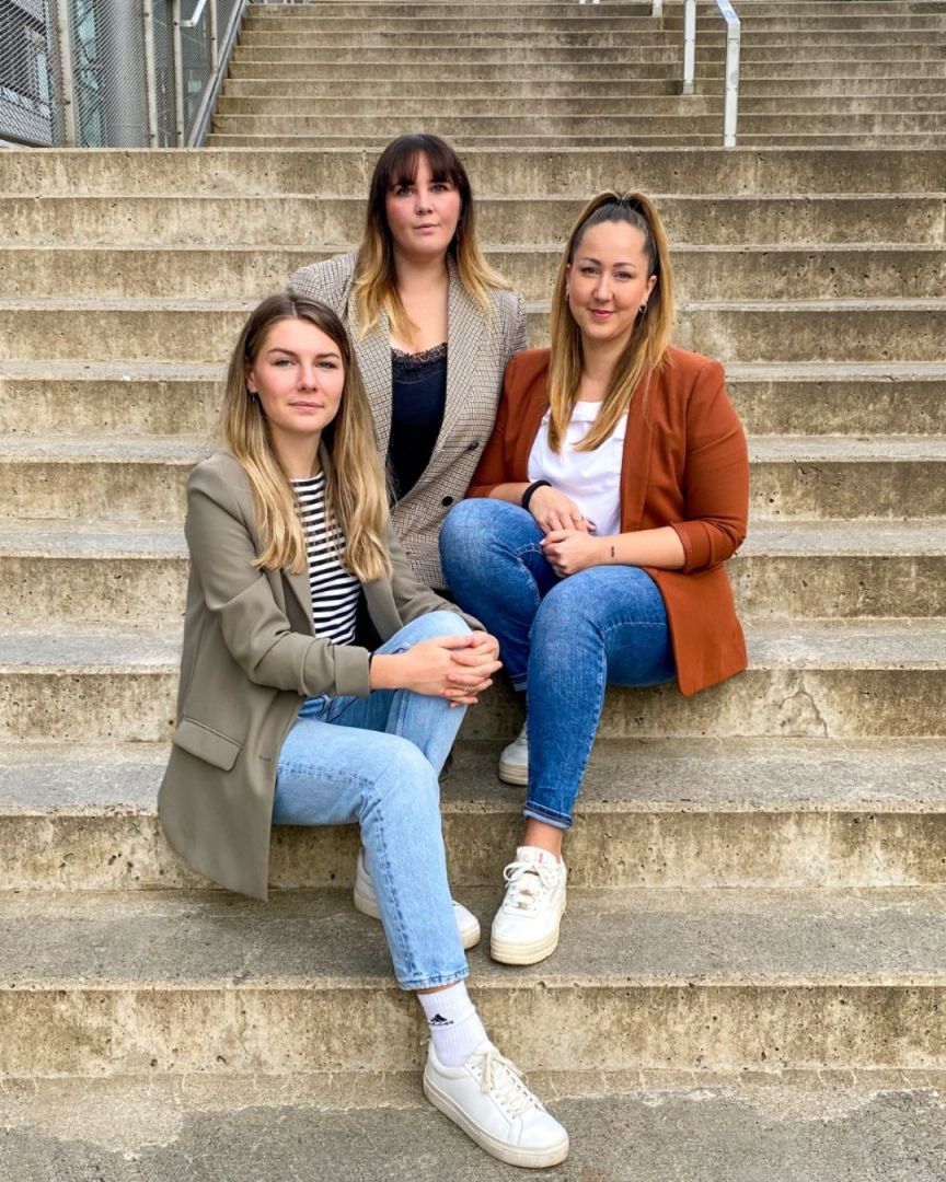 Drei Frauen sitzen auf Betonstufen, tragen Jeans und Blazer, lächeln in die Kamera.