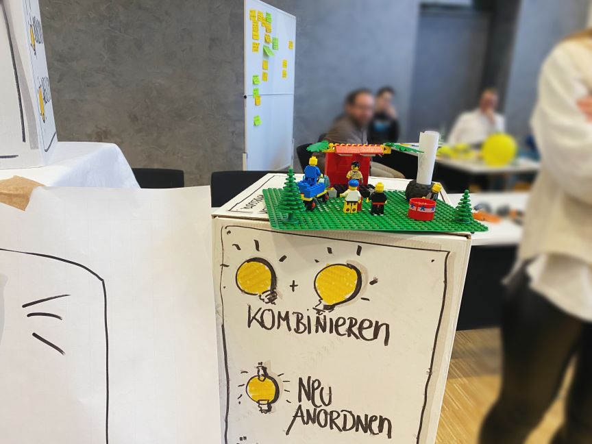 LEGO-Figuren auf grüner Platte, darunter Flipchart mit "Kombinieren" und "Neu Anordnen", Personen im Hintergrund.