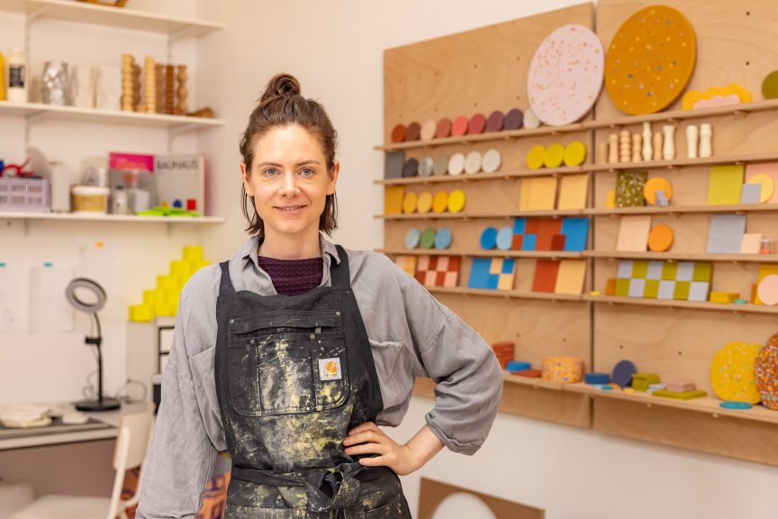 Frau mit Dutt und schmutziger Schürze steht in einem Atelier vor einem Regal mit bunten Keramikmustern.