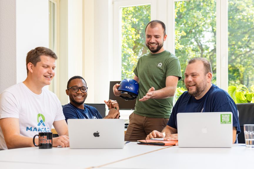 Vier Männer in hellem Büroraum, drei sitzen vor Laptops, einer steht und hält eine blaue Mütze mit Aufschrift „KAJK".