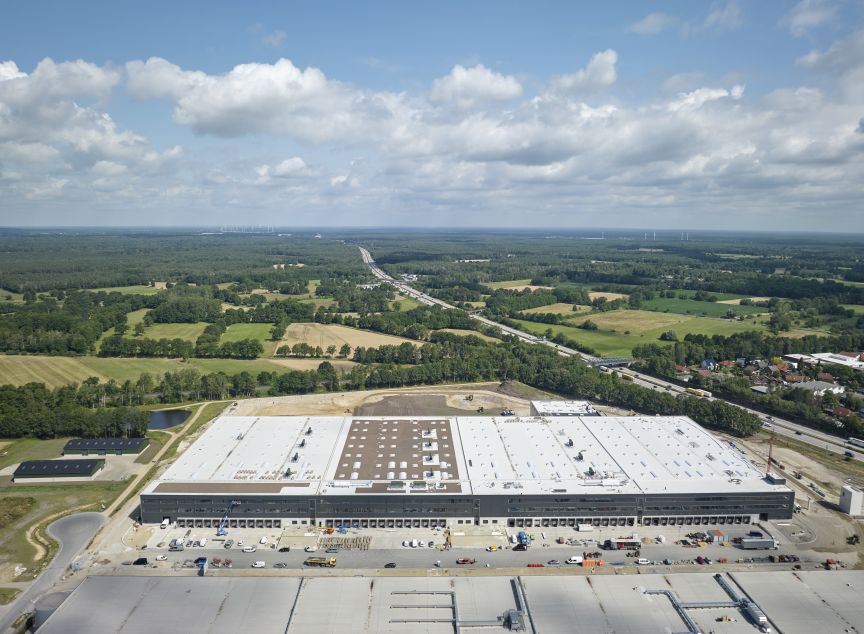 Luftaufnahme eines großen Lagergebäudes mit Laderampen, umgeben von Autobahn, Feldern und Wäldern.
