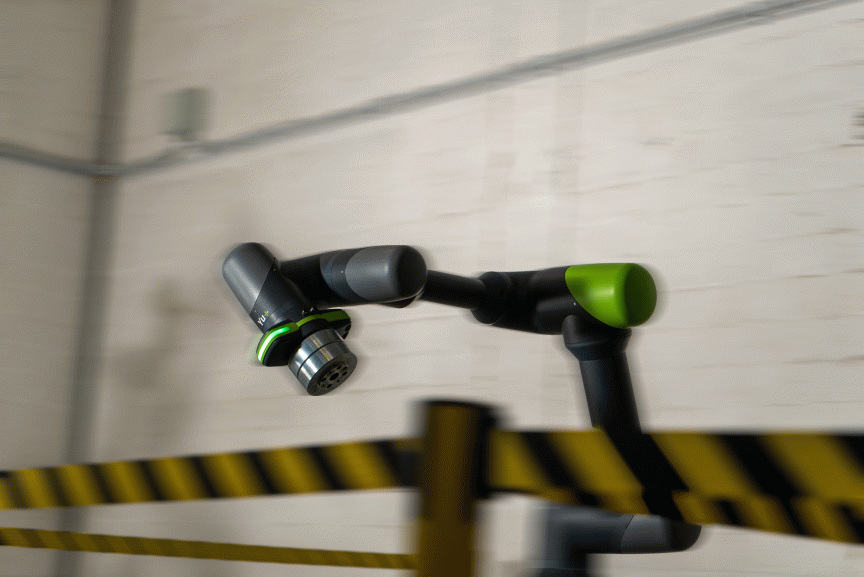 Schwarzer Roboterarm mit grünen Gelenken in Bewegung, montiert an weißer Wand, gelb-schwarzes Absperrband im Vordergrund.
