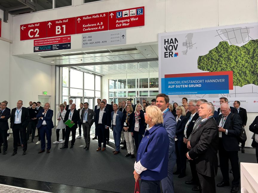 Gruppe von Personen in Messehalle, Hannover-Banner mit Text „Immobilienstandort Hannover auf gutem Grund", Hallenbeschilderung B1, C2.