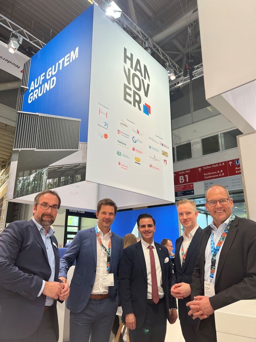 Fünf Männer in Businesskleidung posieren vor einem Hannover-Messestand mit dem Schriftzug „Auf gutem Grund".