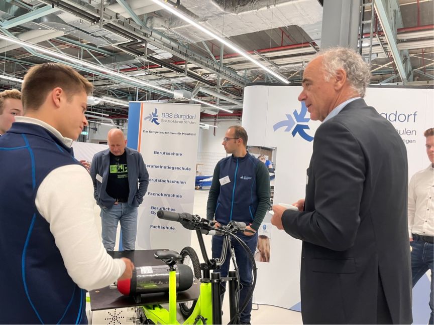Mehrere Personen stehen an einem E-Bike-Stand der BBS Burgdorf Berufsschule auf einer Messe in einer Halle.